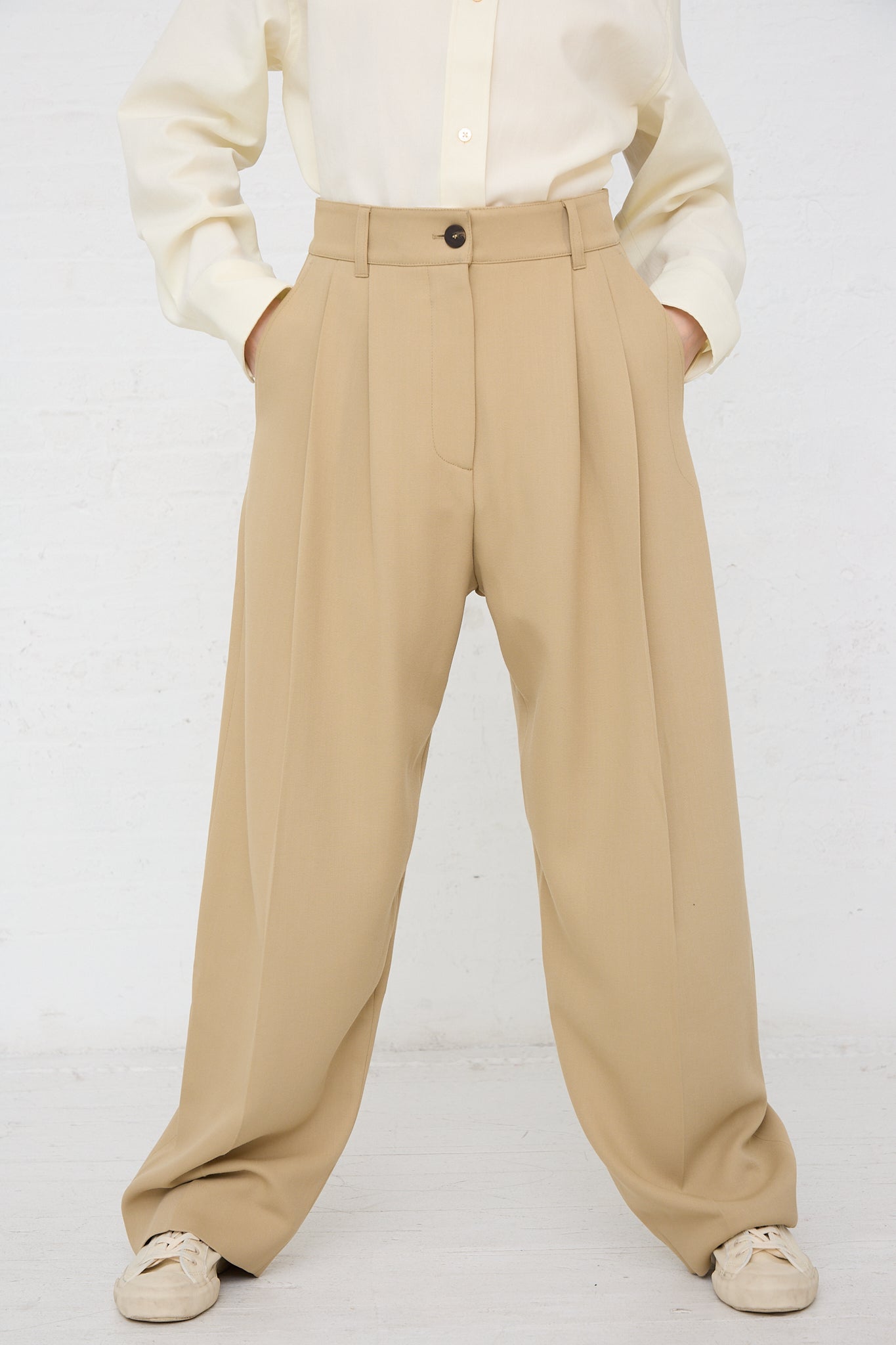 A model in a white shirt and **Acuna Double Pleat Front Trouser in Sand** by Studio Nicholson. Front view. Model's hands are in pockets.