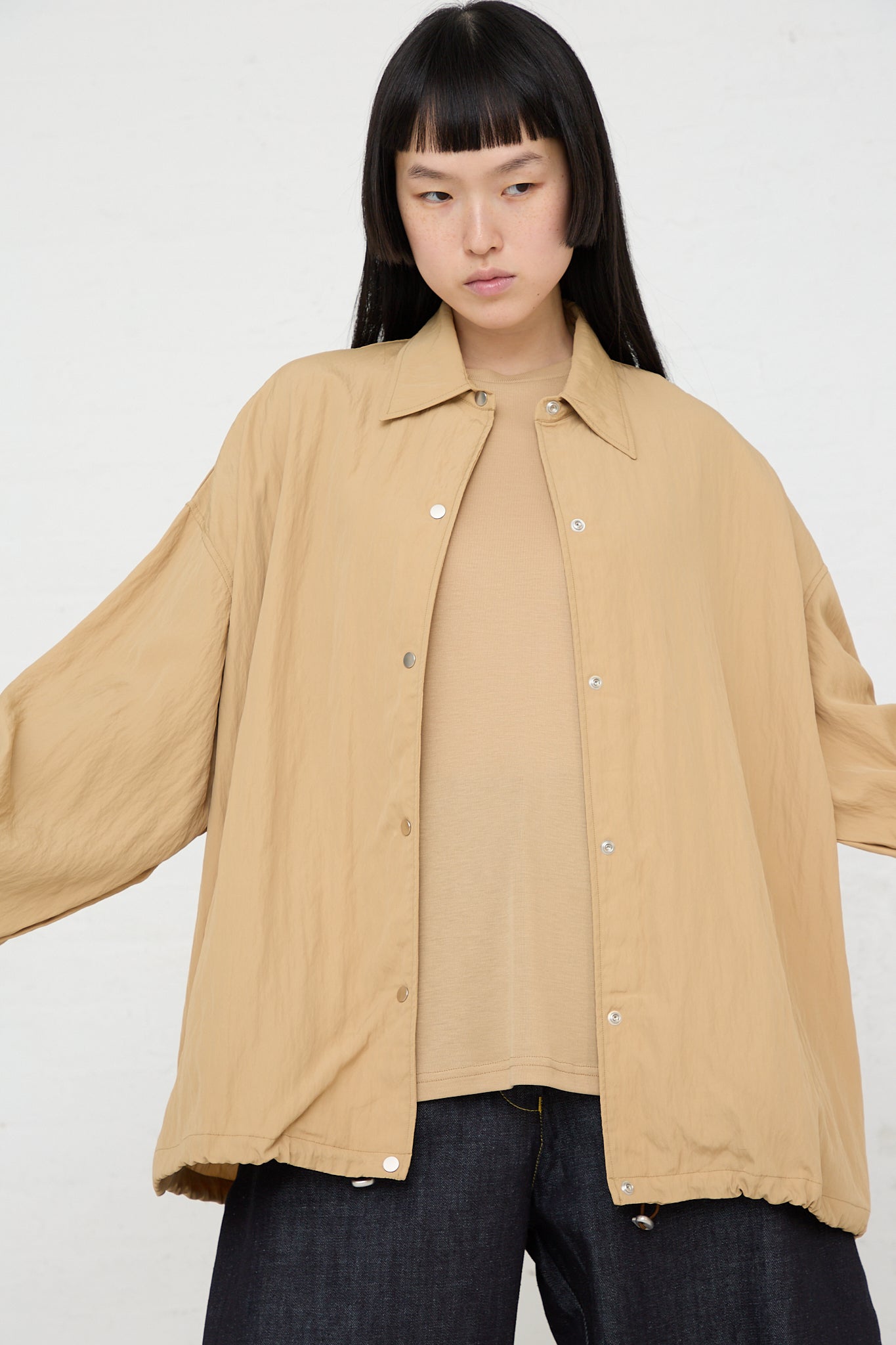 A model wearing a viscose blend tan Sprung Coach Jacket in Sand by Studio Nicholson, with elasticated cuffs, paired with jeans.