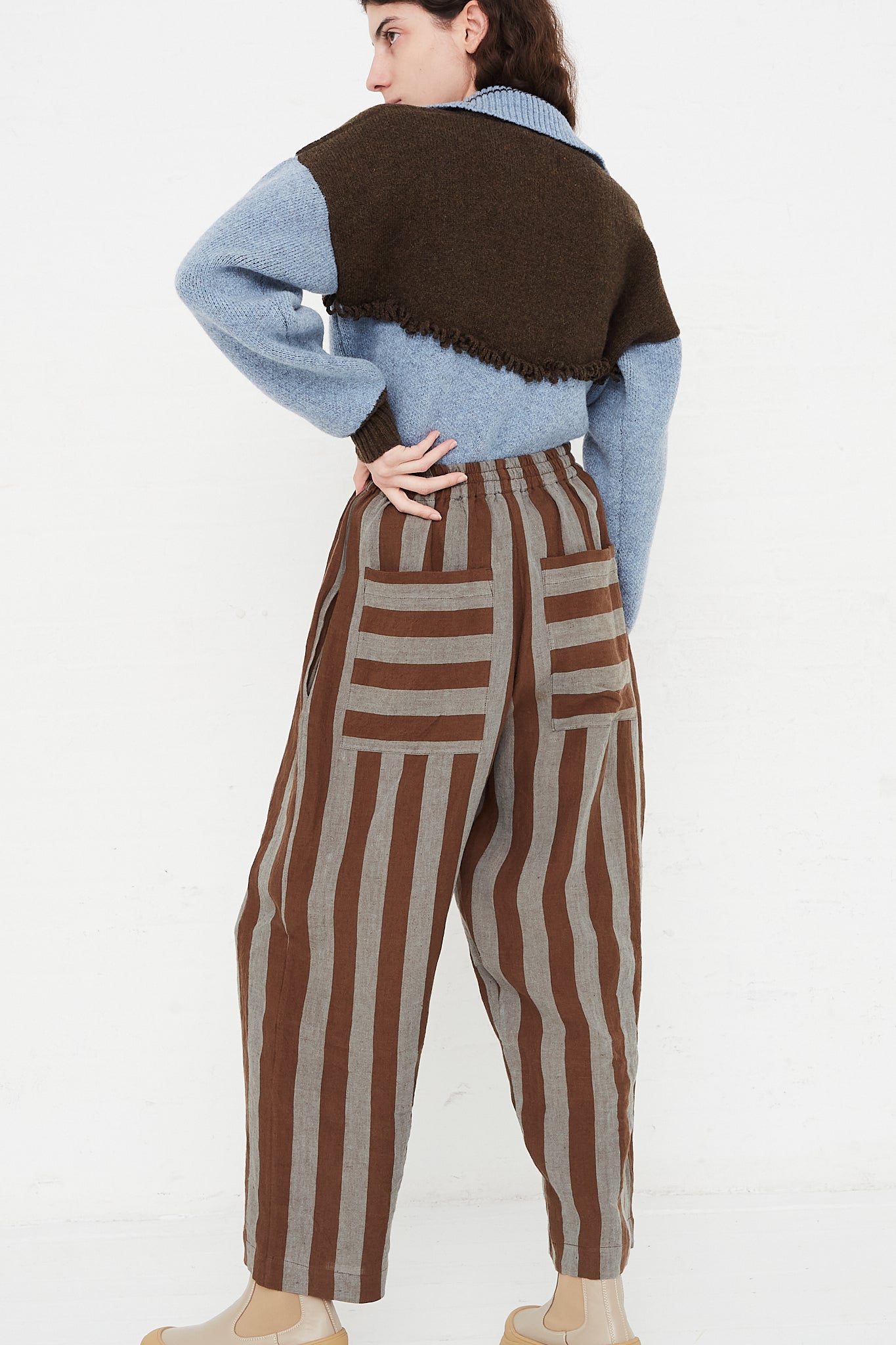 A model wearing a high waist trouser in a striped Irish linen. Brown and blue stripe. Features an elasticated waist and a wide straight leg. Full length and back view of model highlight full pant, sweater and boots. Showing both pocket placements and details. Pockets feature horizontal stripe in contrast to vertical pant stripe. Designed by Cawley - Oroboro Store