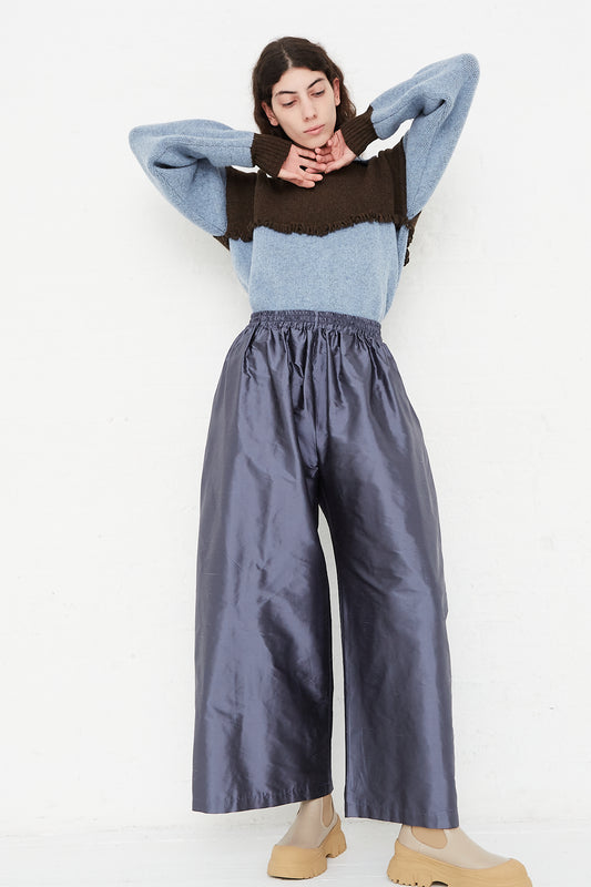 A model wearing a high waist trouser in a shiny silk dupion. Features an elasticated waist and a wide balloon leg.  Color: grey. Full length and full view highlighting the pants, sweater, and boots. Arms raised showing sweater details.  Designed by Cawley - Oroboro Store