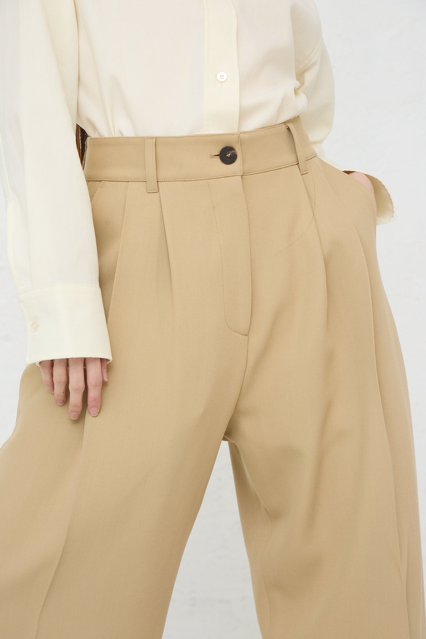 A woman wearing the Studio Nicholson Acuna Double Pleat Front Trouser in Sand and a white shirt. Up close view of front detail.