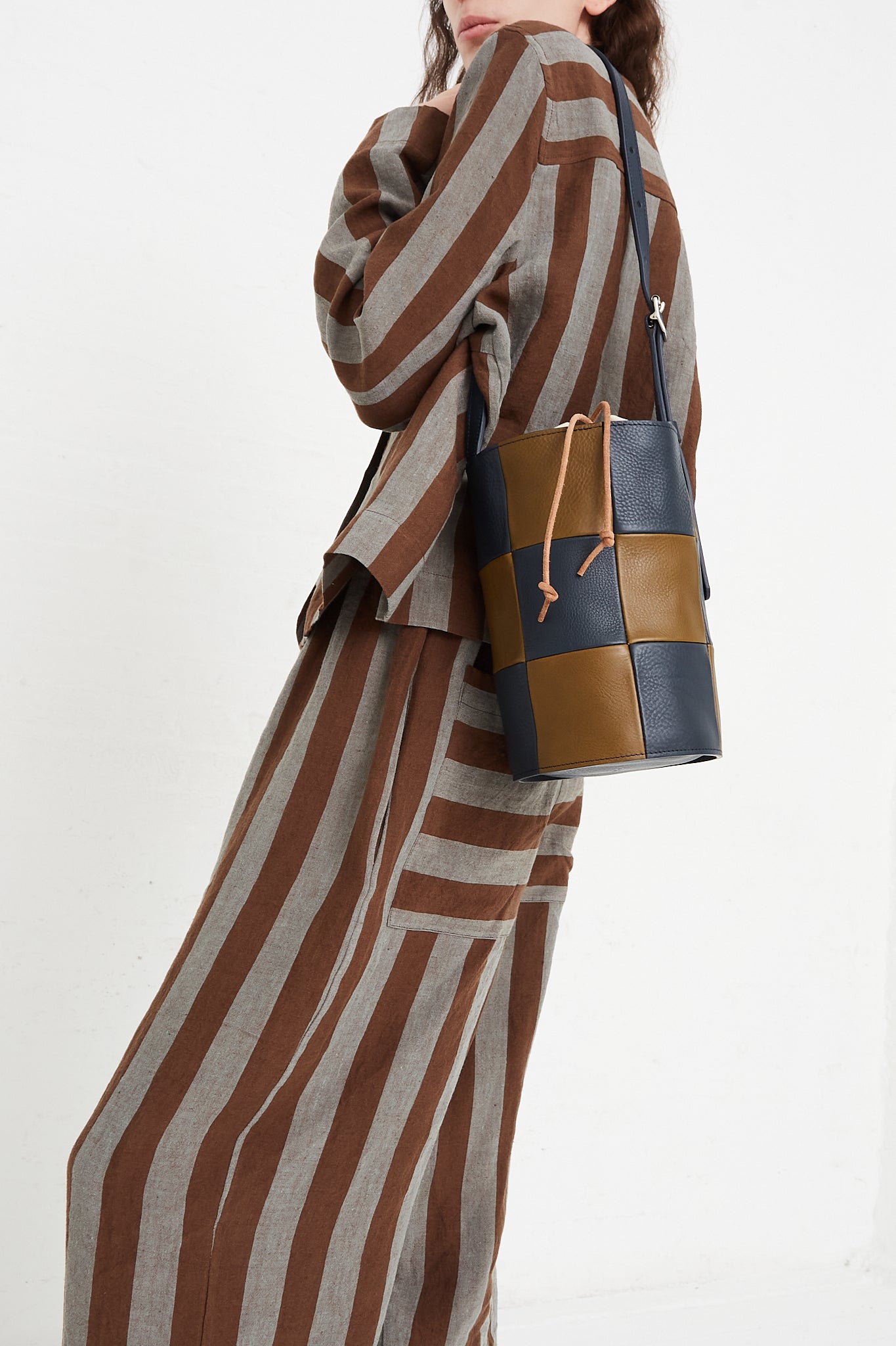 A model holding a bucket bag in a grainy patchwork leather. Features an adjustable strap with silver hardware, and an interior canvas bag with leather tie closure. Side view and upclose. Designed by Cawley - Oroboro Store