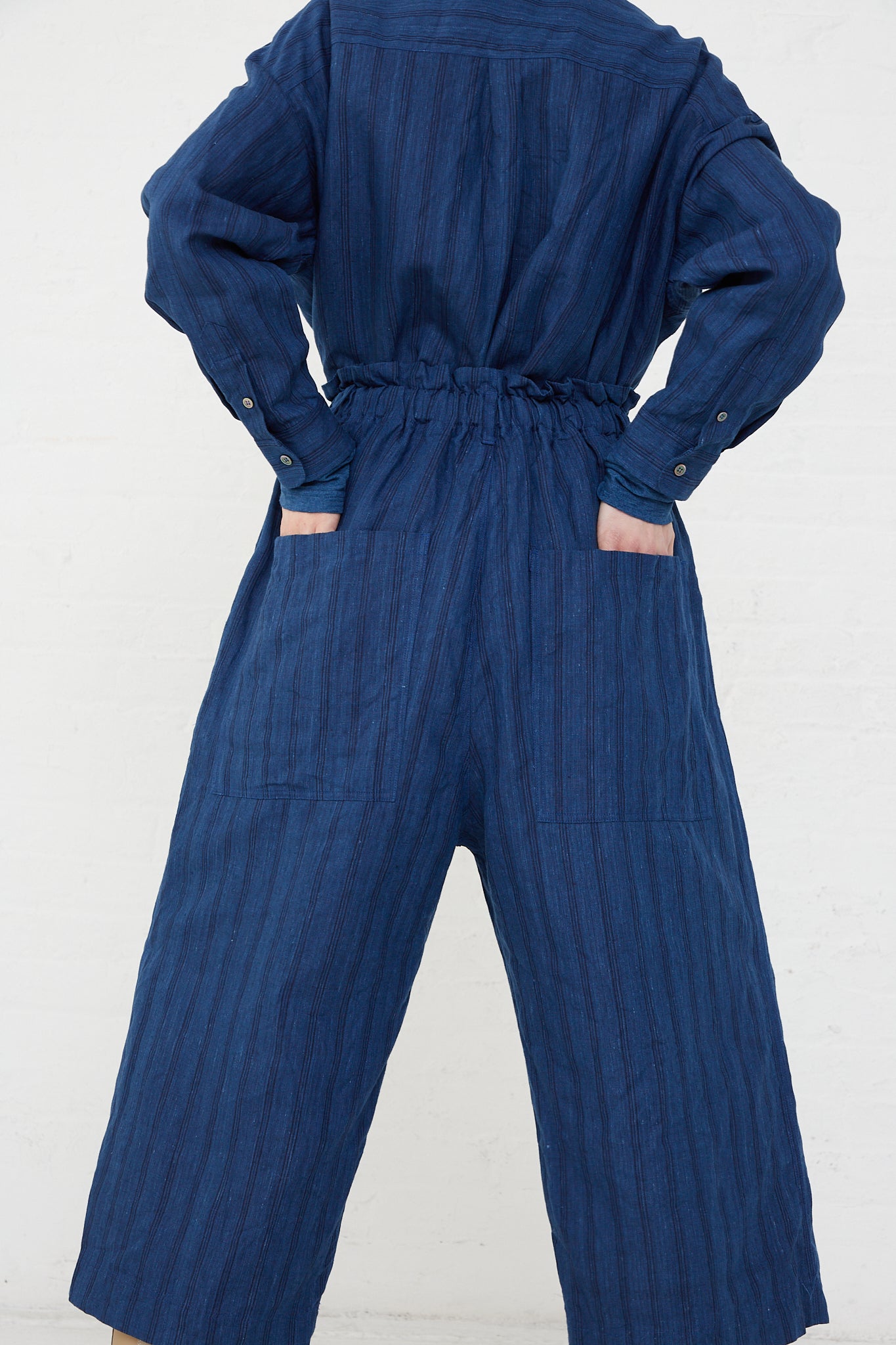 The back view of a woman in a Ichi Antiquités Indigo Stripes woven linen jumpsuit, featuring an elasticated waist and wide leg pant.