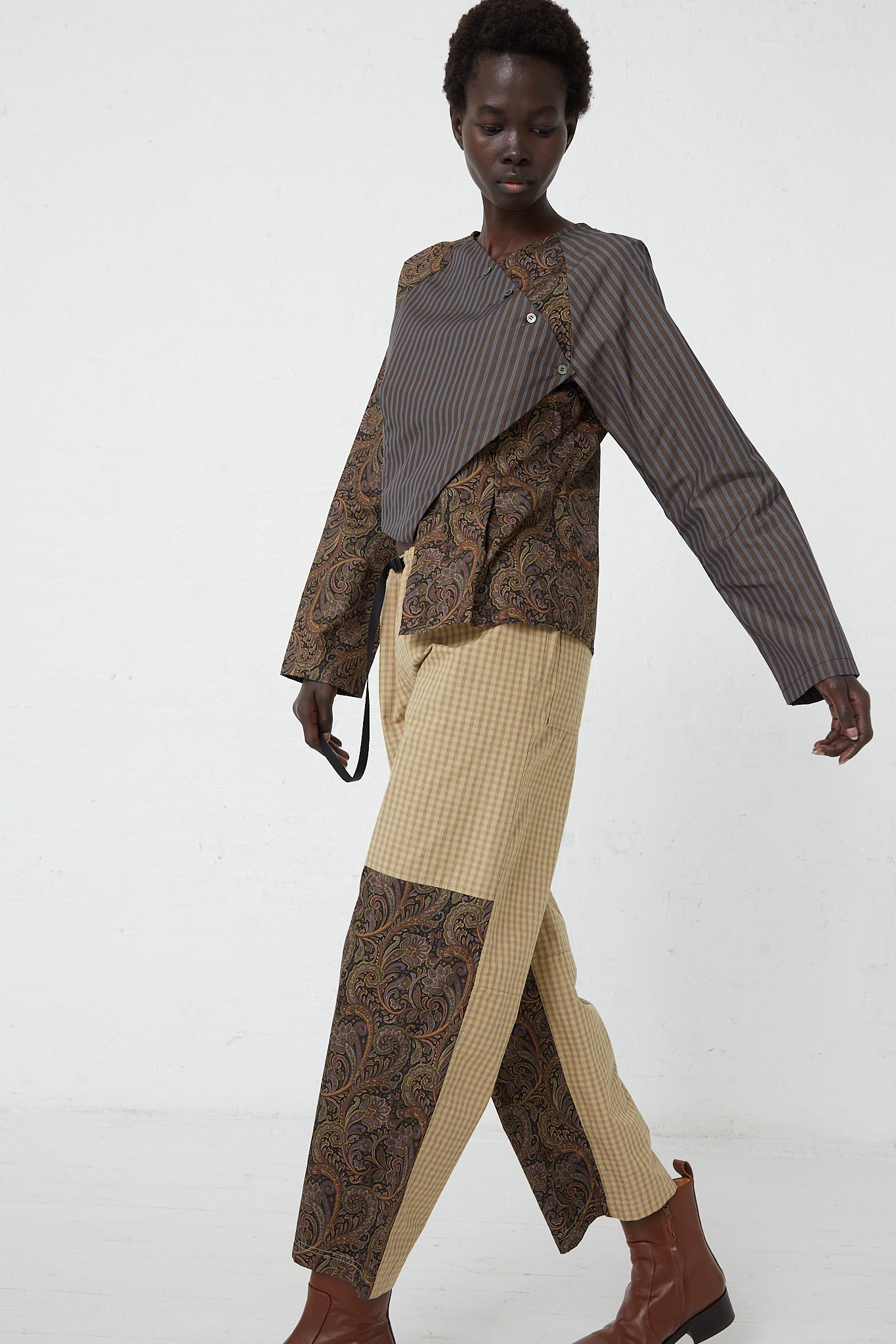 A woman in a brown and tan outfit walking on a white floor, showcasing her SC103 Check Cotton Domain Pant in Tent.