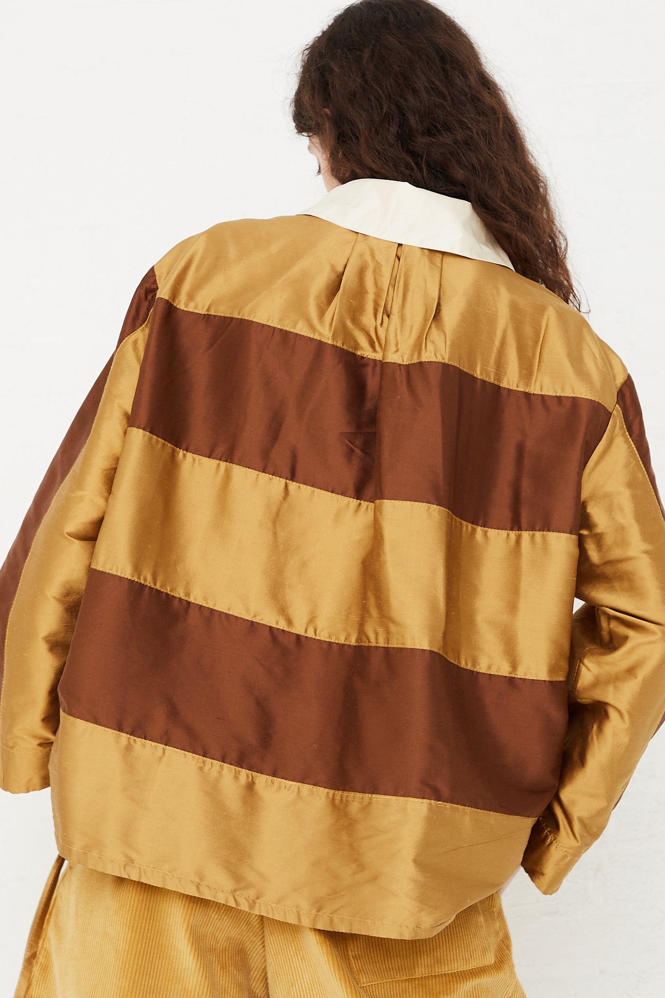 A model wearing a striped rugby shirt in a shiny silk doupion. Features a contrast fold-over collar and button placket, with an adjustable drawstring at hem. Back view and up close. Designed by Cawley - Oroboro Store 