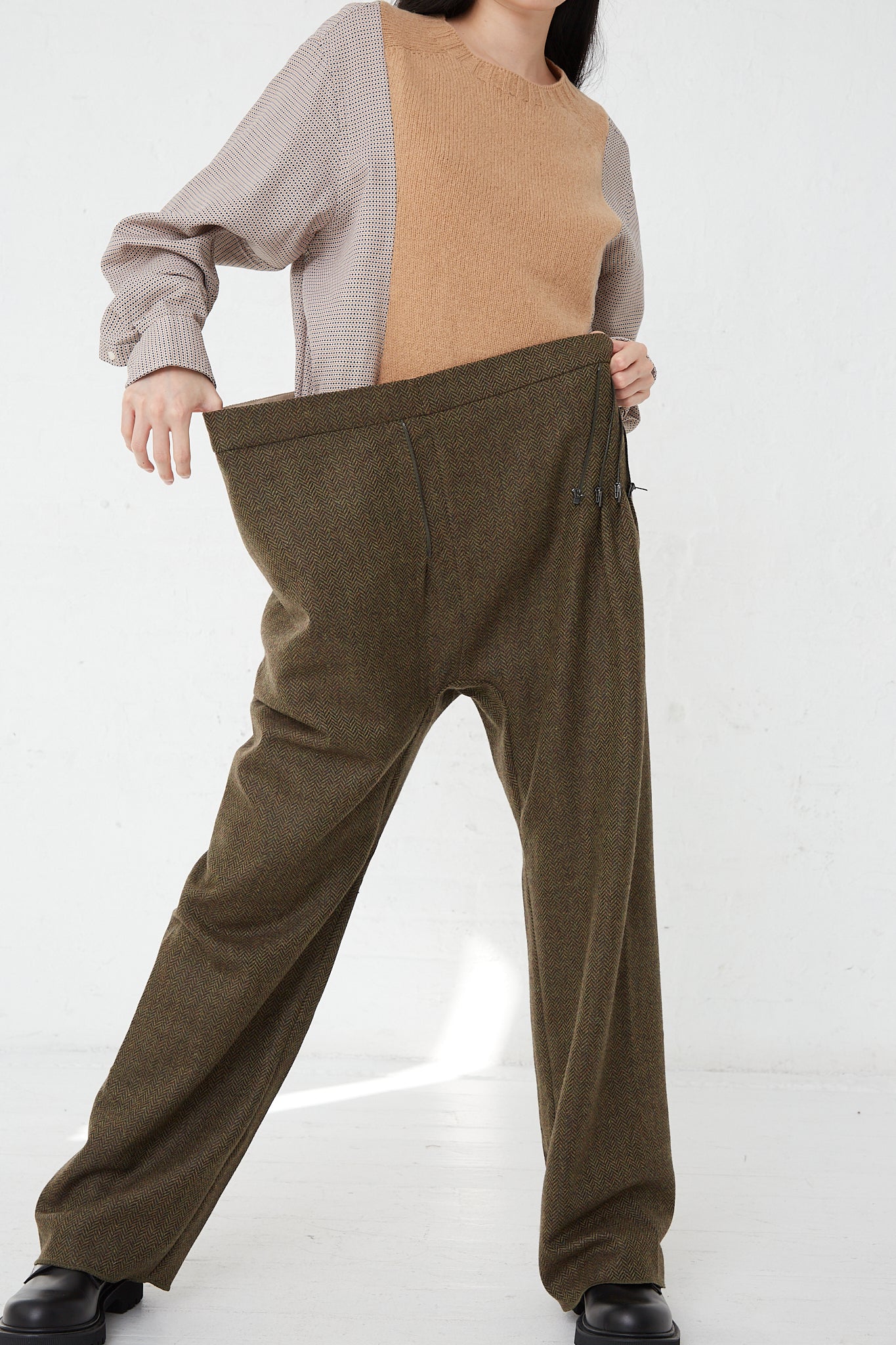 A woman is standing in front of a white wall holding a pair of Bless No. 75 Wool Long Pant in Purple Greenish Tweed with an adjustable zipper accent in front.